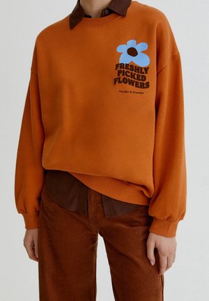 Persona que lleva una sudadera naranja con un gráfico de flor azul y texto ""Freshly Picked Flowers"", combinada con camisa marrón y pantalones de pana.