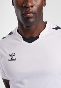Weißes Sporttrikot mit V-Ausschnitt, schwarzen Akzenten an den Schultern und einem Logo auf der Brust. Glatte Textur, leichtes Material.