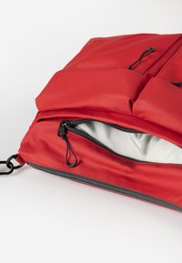 Primo piano di uno zaino rosso in nylon, con una tasca con zip, fodera interna liscia e hardware del cordoncino regolabile.