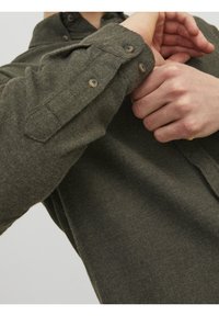 Camicetta verde texturizzata, con bottoni e realizzata in un tessuto spesso. Presenta un colletto e due bottoni visibili sul polsino.