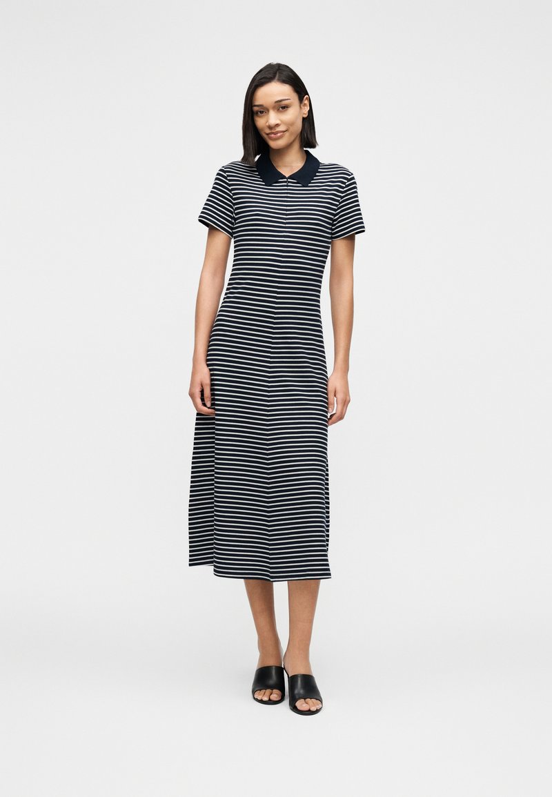 Γυναίκα που στέκεται φορώντας μεσομήκη φόρεμα με οριζόντιες ασπρόμαυρες ρίγες, μαύρο γιακά και μαύρα παπούτσια ανοιχτής μύτης, σε λευκό φόντο.