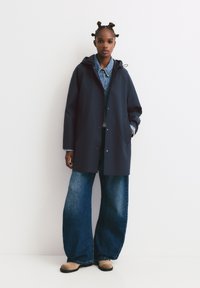 Marineblå vandtæt jakke med hætte, trykknaplukning foran og sidelommer, lagdelt over en denimskjorte og parret med vide jeans.