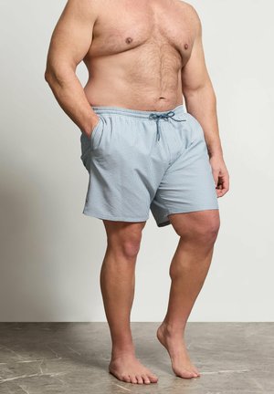 Mężczyzna z gołą klatką piersiową stojący na boso, ubrany w jasnoniebieskie i białe paskowane szorty ze ściągaczem, z rękami w kieszeniach na jednolitym tle.