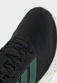 Zapatilla deportiva negra con parte superior texturizada, detalles en verde y suela blanca con salpicaduras. Los cordones son oscuros y gruesos para un ajuste seguro.