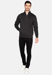 Felpa nera a zip realizzata in materiale morbido, con colletto alto e polsini a coste, abbinata a pantaloni neri slim-fit e sneakers bianche.