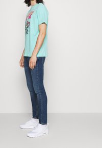 Camiseta gráfica turquesa, tela de algodón ligera, cuello redondo. Combinada con vaqueros ajustados azul oscuro y zapatillas deportivas blancas.