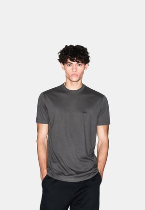 T-shirt grigia a maniche corte con scollo rotondo, realizzata in tessuto liscio, con un piccolo logo nero sul lato sinistro del petto, abbinata a pantaloni scuri.