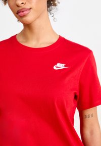 Tricou roșu din bumbac, cu mâneci scurte și guler rotund. Prezintă logo-ul alb Nike pe partea stângă a pieptului. Textură netedă, design ajustat.