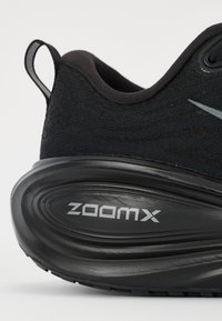 Svarta sportskor med ett strukturerat tygöverkåpa och en vadderad, böjd svart sula. "ZOOMX"-logotyp i silver på sidan.