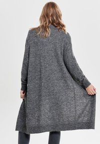 Vrouw met lichtbruin haar tot op de schouders, gekleed in een lange, losse, grijze gebreide cardigan, die de zijkanten van haar lichaam afhoudt.