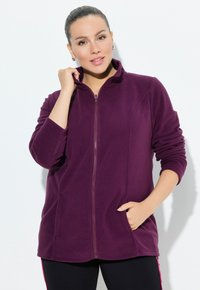 Donna che indossa una giacca in pile viola con zip, con una mano in tasca e l'altra che sistemando il colletto, su uno sfondo chiaro e uniforme.