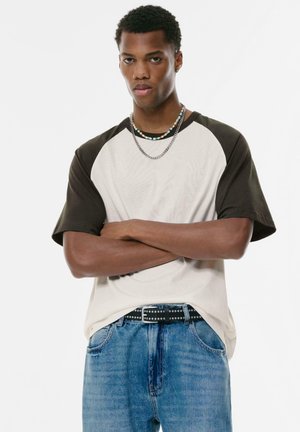 T-shirt med raglanærmer i cremefarvet krop og mørkebrune ærmer. Blødt bomuldsstof, rund halsudskæring, parret med blå denim jeans og et nitted bælte.