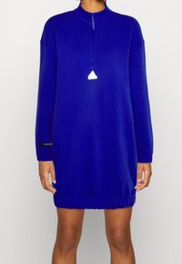 En koboltblå långärmad sweatshirtklänning med halvzip-nacke och elastisk fåll, prydd med en vit logodetalj på bröstet.