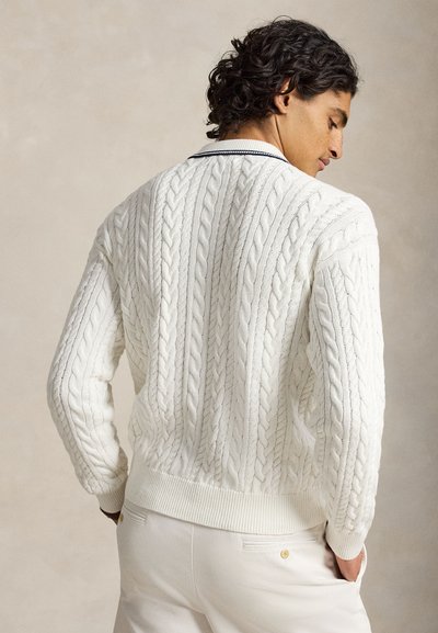 Homme aux cheveux bouclés portant un pull en maille torsadée blanc et un pantalon blanc, debout les mains dans les poches sur un fond neutre.