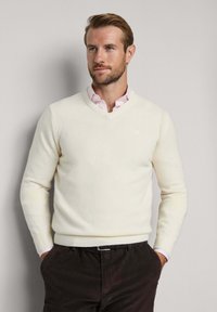 V-Ausschnitt-Pullover aus cremefarbenem Strickstoff, mit gerippten Bündchen und Saum. Getragen über einem hellrosa Hemd mit Kragen, kombiniert mit dunklen Hosen.