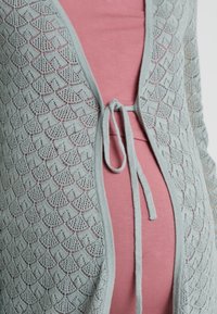 Cardigan léger en tricot bleu clair avec un motif de diamant texturé et un lien à l'avant, porté sur un haut rose. Tissu doux, design ouvert.