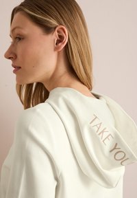 Sudadera blanca con textura acanalada, que presenta un mensaje bordado "Llévate" en la capucha. Tejido suave y corte relajado.