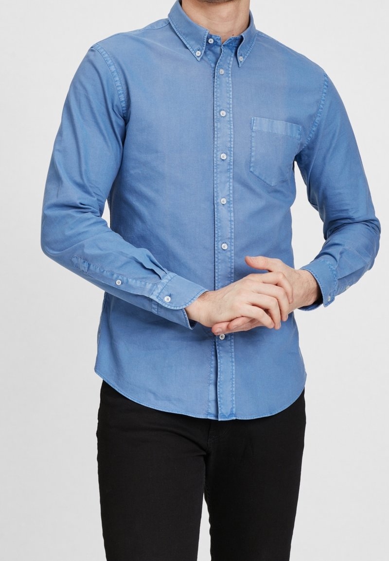 Homme portant une chemise bleue à boutons avec une poche poitrine et un pantalon noir, les mains jointes devant, debout devant un fond uni.