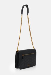 Sort quiltet håndtaske med en guldkæde som rem, med en lynlåslomme foran og mærket "GUESS" præget nederst.