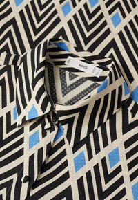 Camisa con un patrón geométrico en negro, crema y azul; incluye collar y detalles de botones. La tela tiene una textura suave.