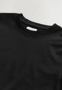 Camiseta de algodón negra con cuello redondo, que presenta una textura suave y una etiqueta denominada "NEXT SIGNATURE" en el interior del cuello.