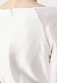 Haut blanc avec une finition en soie lisse, présentant une fermeture éclair discrète à l'arrière et un col arrondi. Texture douce avec des coutures minimales.