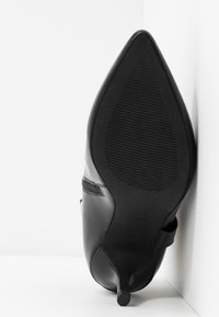 Zapato de tacón negro con puntera afilada, parte superior de cuero liso, suela de goma texturizada y un elegante detalle de tira en el lateral.