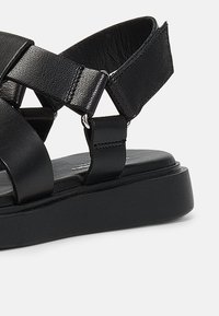 Sorte lær sandaler med kryssende remmer, flat såle og justerbare ankelremmer. Glatt tekstur med metallaksenter.