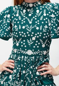 Teal mekko, jossa on valkoista kukkakirjontaa, puff-hihat, korkea kaulus ja kerätty hameen yksityiskohta. Kuvioina on erilaisia kukkia.