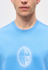 Άνδρας που φοράει γαλάζιο T-shirt με στρογγυλή λαιμόκοψη και λευκό κυκλικό λογότυπο και γραφικό στο στήθος, ορατό από τον λαιμό μέχρι τον άνω κορμό.