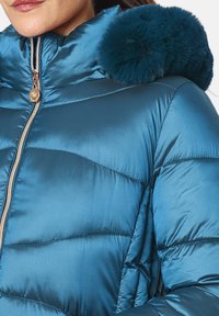 Chaqueta de plumas color teal con un cuello alto y un acento de pom-pom de piel. La tela es brillante, con secciones acolchadas y un zipper dorado.