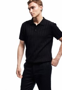 Junger Mann mit heller Haut und kurzen Haaren, der ein schwarzes kurzärmliges Strick-Polohemd und schwarze Hosen trägt, und vor einem weißen Hintergrund steht.