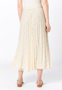 Krämfärgad spetsfållad kjol med en slät midja, med blommönster och en flytande design, ihop med ljusbruna platta sandaler.