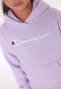 Levandulová mikina s kapucí, která má přední kapsu, vyšívané logo "Champion" v bílé barvě a malé červené akcenty v designu loga.