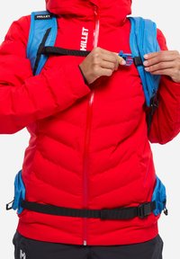 La veste isolante rouge présente une fermeture éclair sur le devant, des sangles ajustables bleues et des boucles noires. La veste a une texture matelassée horizontale.