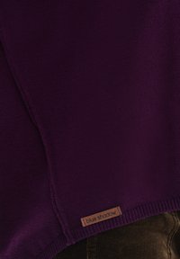 Fioletowy sweter dzianinowy z prążkowanym dołem i drewnianą metką z logo "blue shadow". Tekstura wydaje się miękka z drobnym splotem.