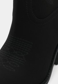 Botín negro con una textura mate suave, que presenta un patrón decorativo embossado y costuras en contraste a lo largo de la suela y los bordes.
