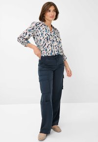 Blouse in lichtroze met een bladpatroon, met een knoopsluiting en een zachte kraag. Donker marine cargo-broek met meerdere zakken. Lichtbeige schoenen.