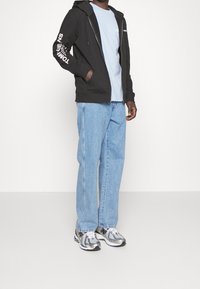 Sweat-shirt zippé noir avec logo blanc sur la manche, t-shirt bleu clair, jean bleu clair et chaussures de sport argentées avec accents noirs.