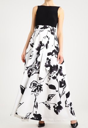 Kvinde iført en ærmeløs sort top og et langt hvidt skørt med store sorte blomsterprint og et matchende stofbælte.