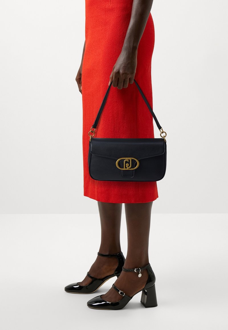Personne portant une robe rouge tenant un sac à main noir avec une fermeture dorée, assorti à des chaussures à talons blocs en cuir verni noir avec des brides à la cheville.