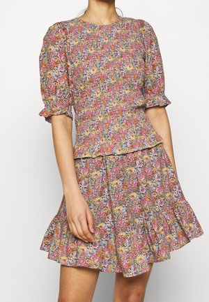 Femme portant une robe courte ajustée à manches bouffantes, avec un corsage smocké et une jupe à ourlet volanté, dans des tons roses, jaunes et bleus.