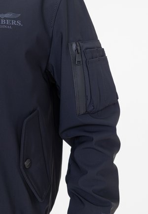 Marineblauer Bomberjacke aus glattem Stoff, mit einer zippten Tasche am linken Ärmel und einem Druckknopf auf der linken Seite.