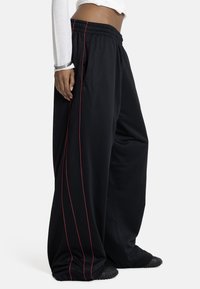 Person trägt lose schwarze Hosen mit roten vertikalen Streifen, weißes cropped Langarmshirt, silberne Armbänder und schwarze strukturierte Schuhe.