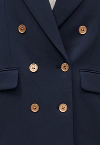 Marineblå dobbeltradet blazer med seks guldfarvede knapper og klaplommer på hver side.