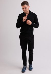 Schwarzer Strickpullover mit einem Reißverschlusskragen, kombiniert mit schwarzen Hosen und blauen Sportschuhen. Schlichte Textur; modernes, tailliertes Design.