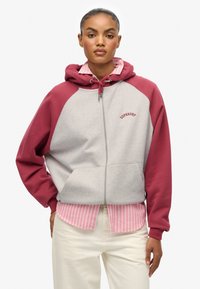Sudadera gris con cremallera, mangas raglán burdeos y cordón. Presenta un bolsillo frontal y el logotipo "SUPERDRY" en texto curvado rojo.
