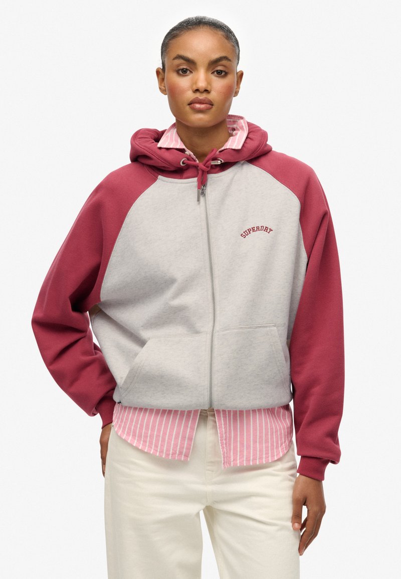 Sudadera gris con cremallera, mangas raglán burdeos y cordón. Presenta un bolsillo frontal y el logotipo "SUPERDRY" en texto curvado rojo.