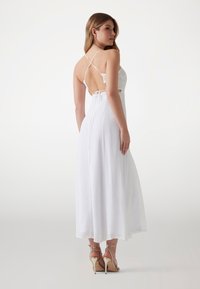 White maxi dress with a low back, lace detailing, thin crisscross straps, and a flowing skirt; paired with strappy, nude high-heeled sandals.