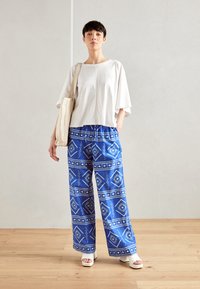 Haut oversize blanc avec des manches tombantes, associé à un pantalon large à motifs bleus présentant des designs géométriques et des sandales compensées blanches.